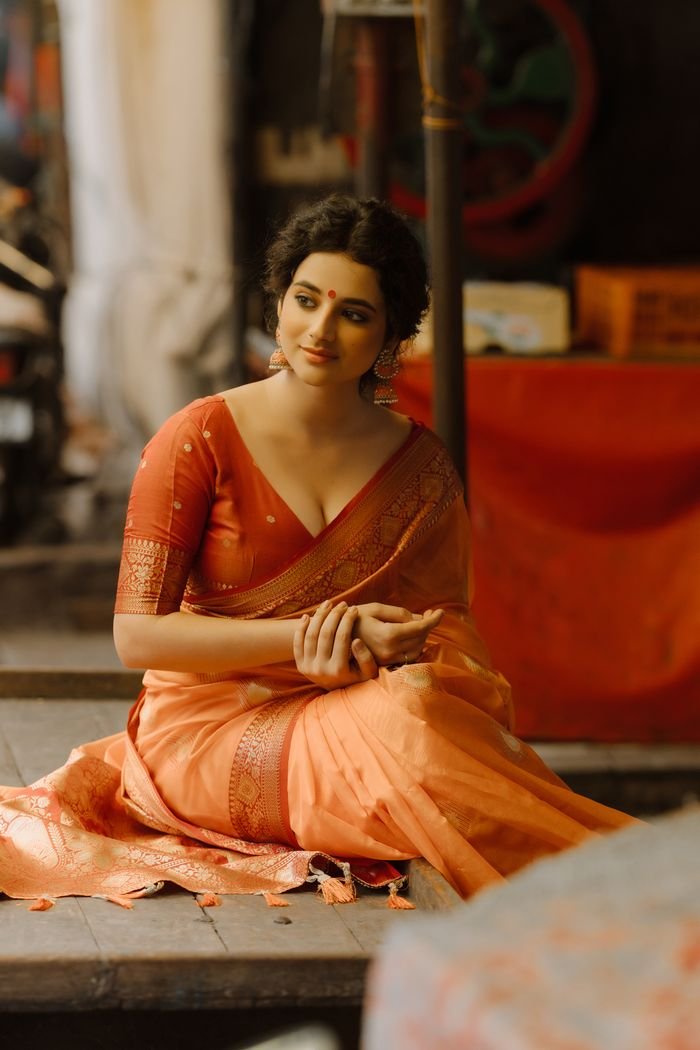 A woman in a traditional orange sari, sitting gracefully indoors with a serene expression.
